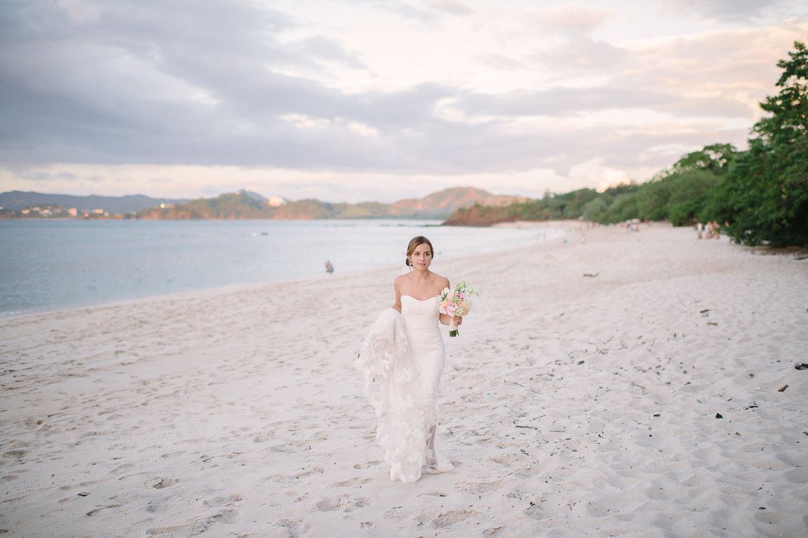 A bride walking along a beach at sunset, holding her train
