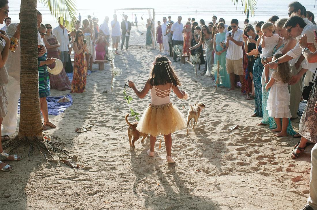 A flower girl leading two dogs down a beach aisle