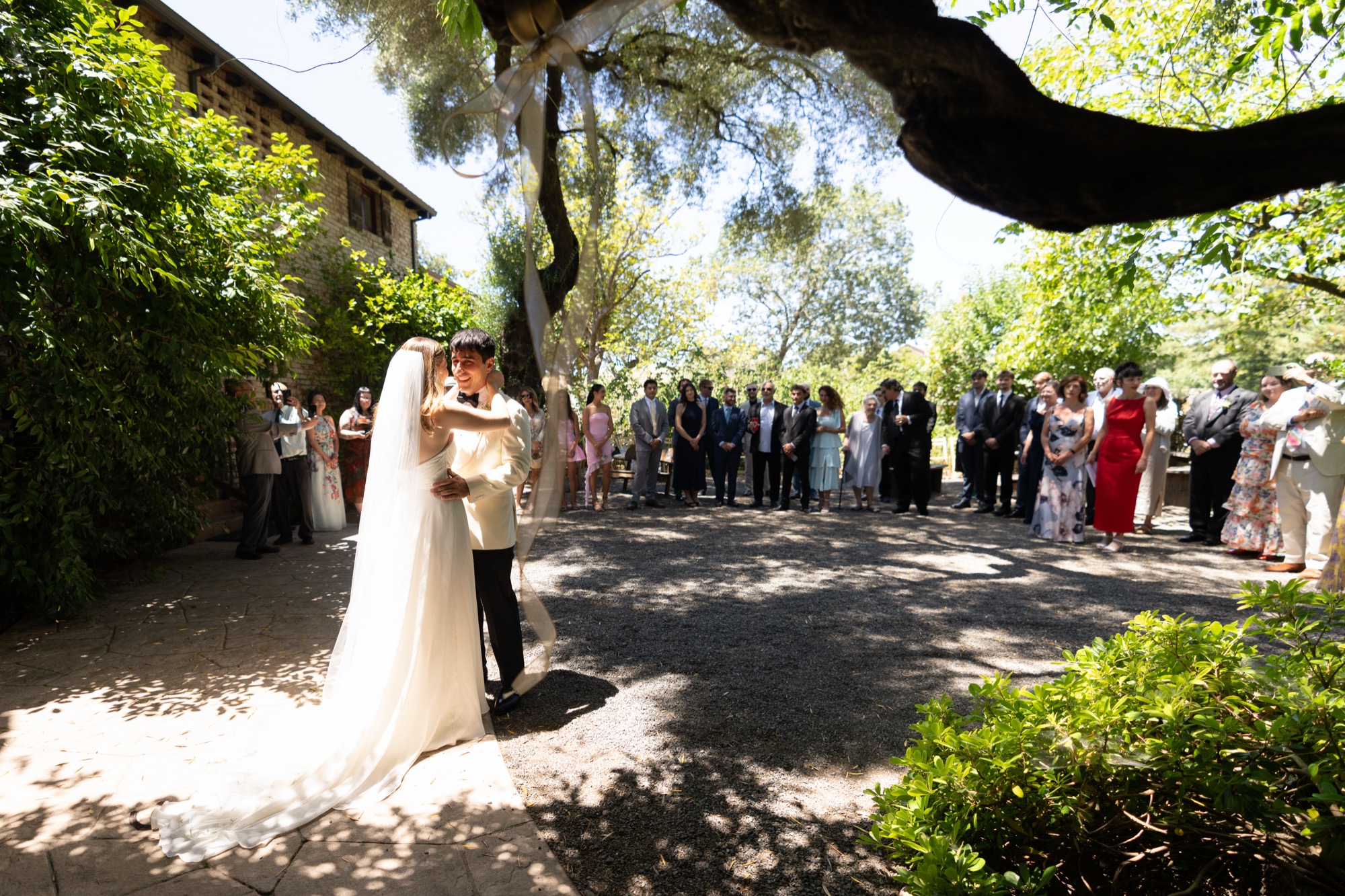 The couple embracing on the drive, guests watching from the shade