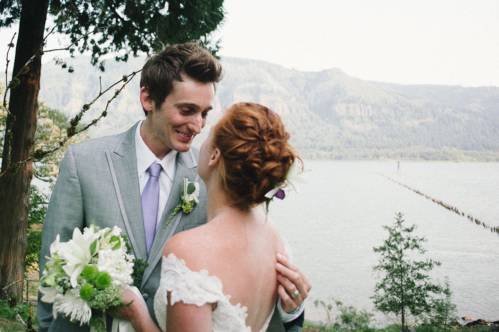 A groom laughing with his bride on a lawn above the river